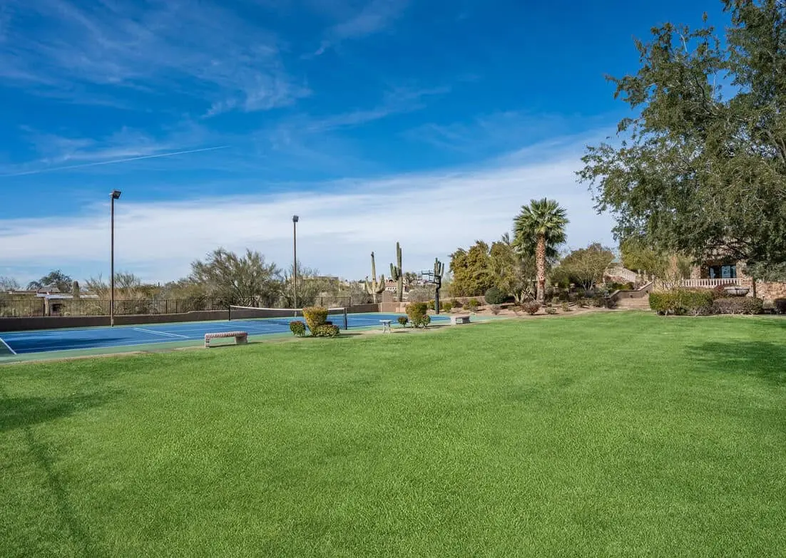 A tennis court sits next to a well-maintained green lawn, enhanced by professional turf installation, with trees, shrubs, and cacti in the background under a bright blue Fountain Hills AZ sky with wispy clouds.