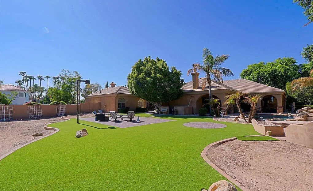 A spacious backyard featuring professional turf installation with artificial turf, desert landscaping, a seating area with chairs and fire pit, palm trees, and a large house in the background beneath a clear blue sky.