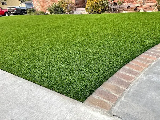 A neatly trimmed, lush green Phoenix artificial grass lawn bordered by a curved brick edging next to a concrete sidewalk, in front of a house with some shrubs and a parked car visible in the background.
