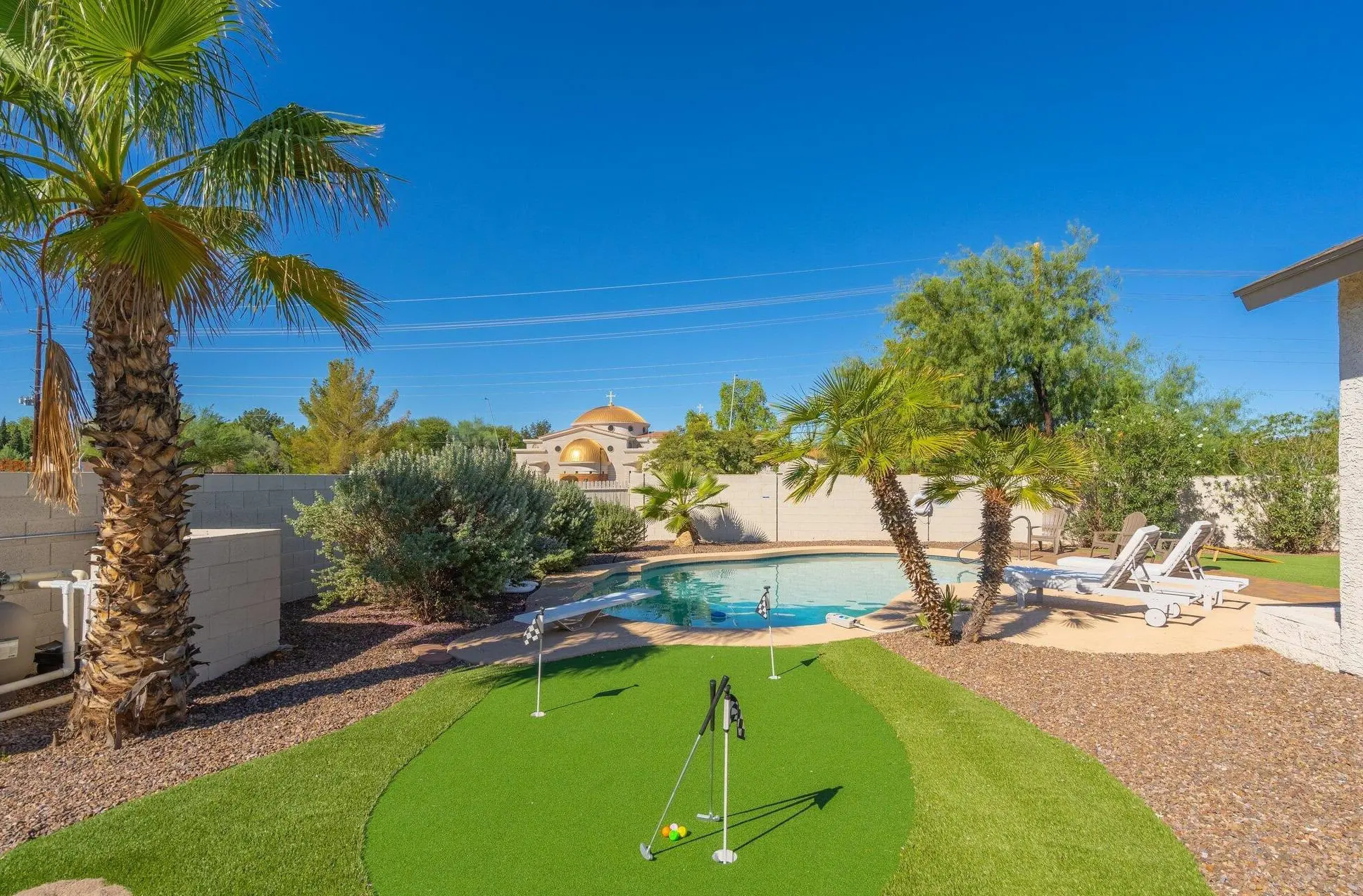 A backyard with professional turf installation, a putting green, palm trees, and landscaping, featuring a curved swimming pool with lounge chairs under a clear blue sky.