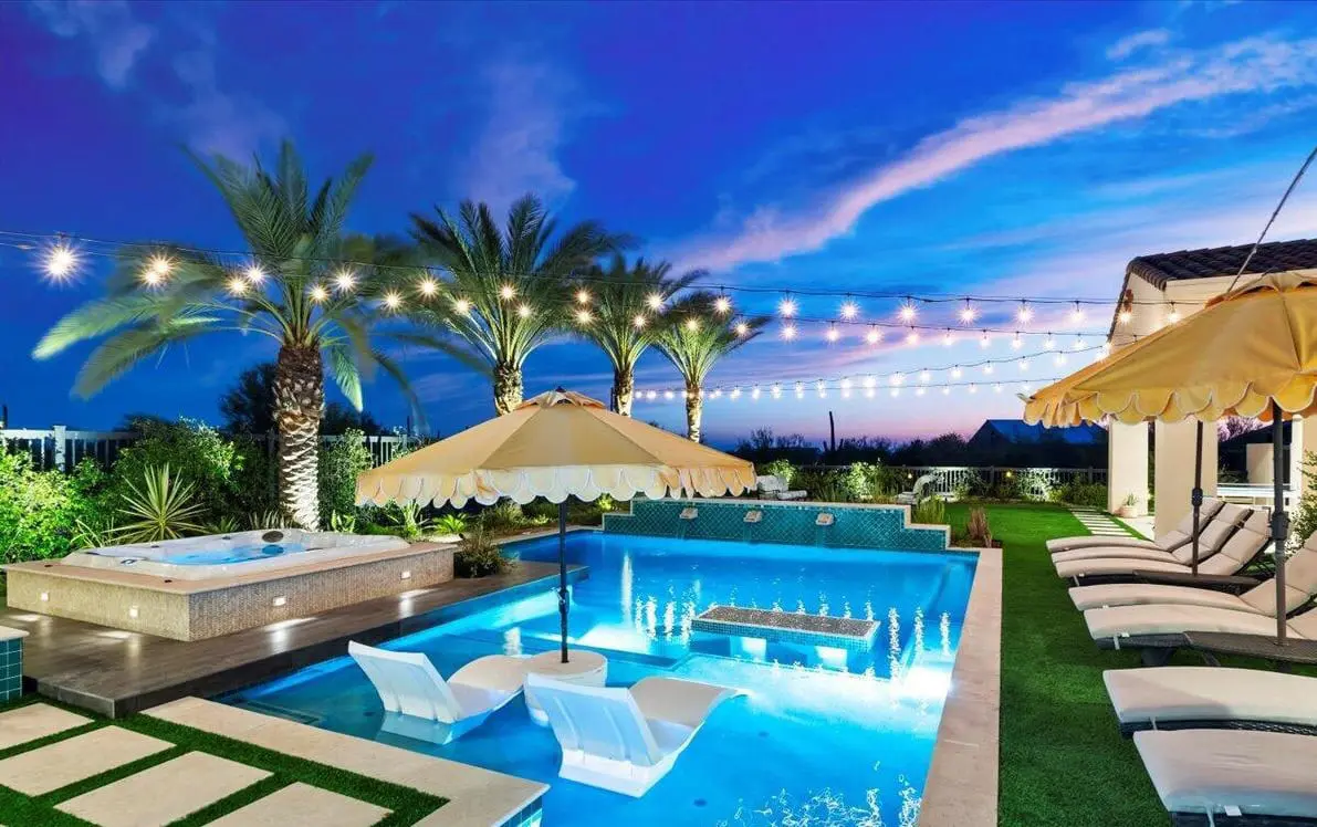 A nighttime view of a Scottsdale, AZ backyard showcases a lit swimming pool surrounded by palm trees. Soft ambient lighting highlights the elegant hardscape design, with outdoor furniture featuring plush cushions. The dark sky enhances the tranquil atmosphere crafted by skilled hardscape contractors.