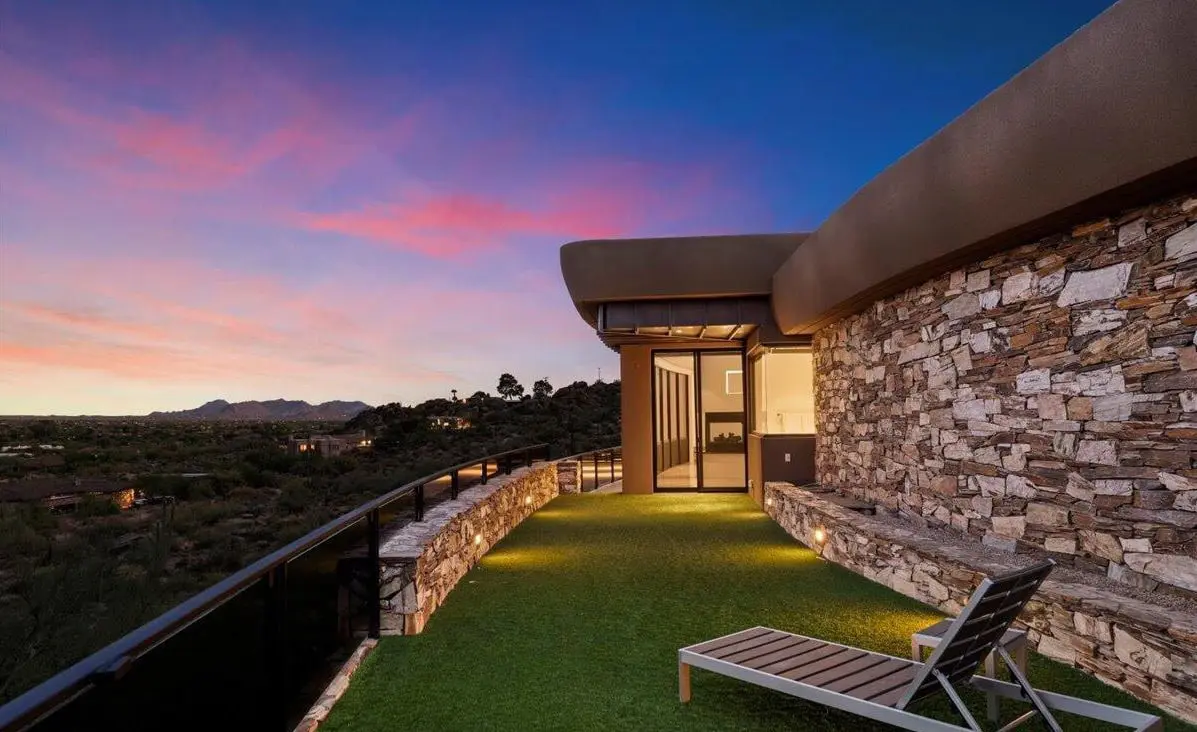 A modern stone terrace with artificial grass, lounge chair, and glass railing overlooks a scenic landscape at sunset.