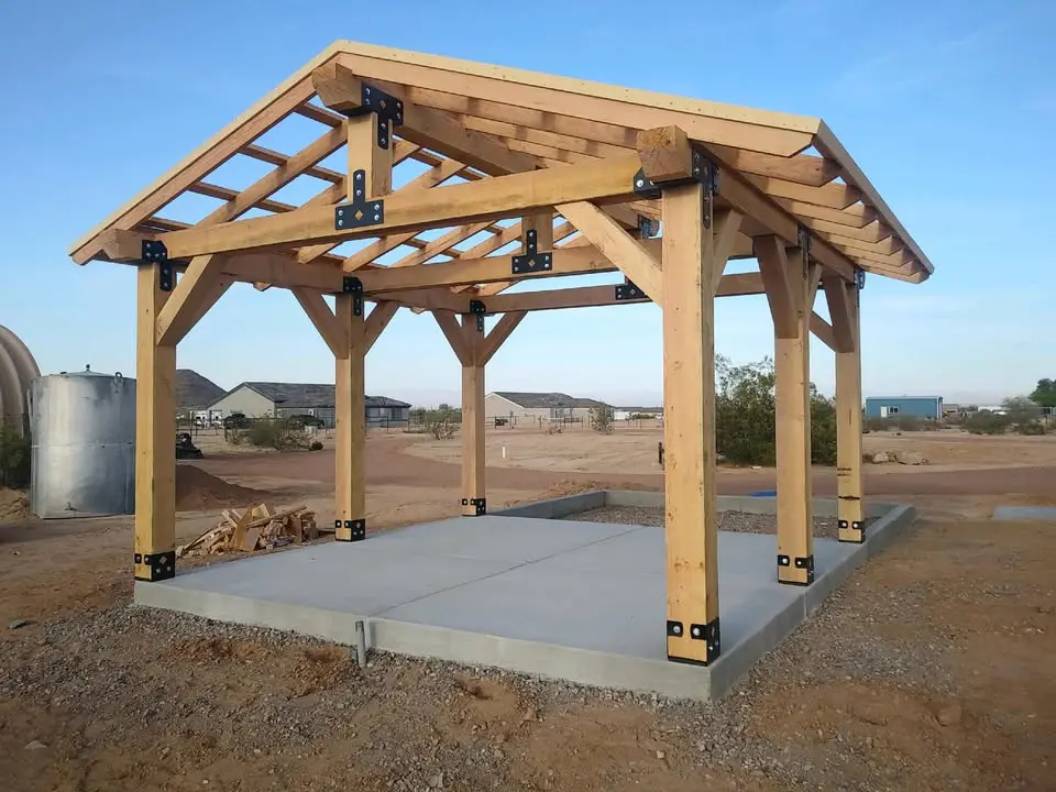 Partially constructed wooden pavilion with exposed timber frame and metal brackets, standing on a concrete foundation in a desert-like outdoor setting. The open-frame structure features a gable roof design without roofing material, supported by thick vertical wooden posts anchored to the concrete base. Surrounding area includes scattered desert plants, dirt ground, nearby houses, and a cylindrical metal water tank. Clear blue sky in the background.