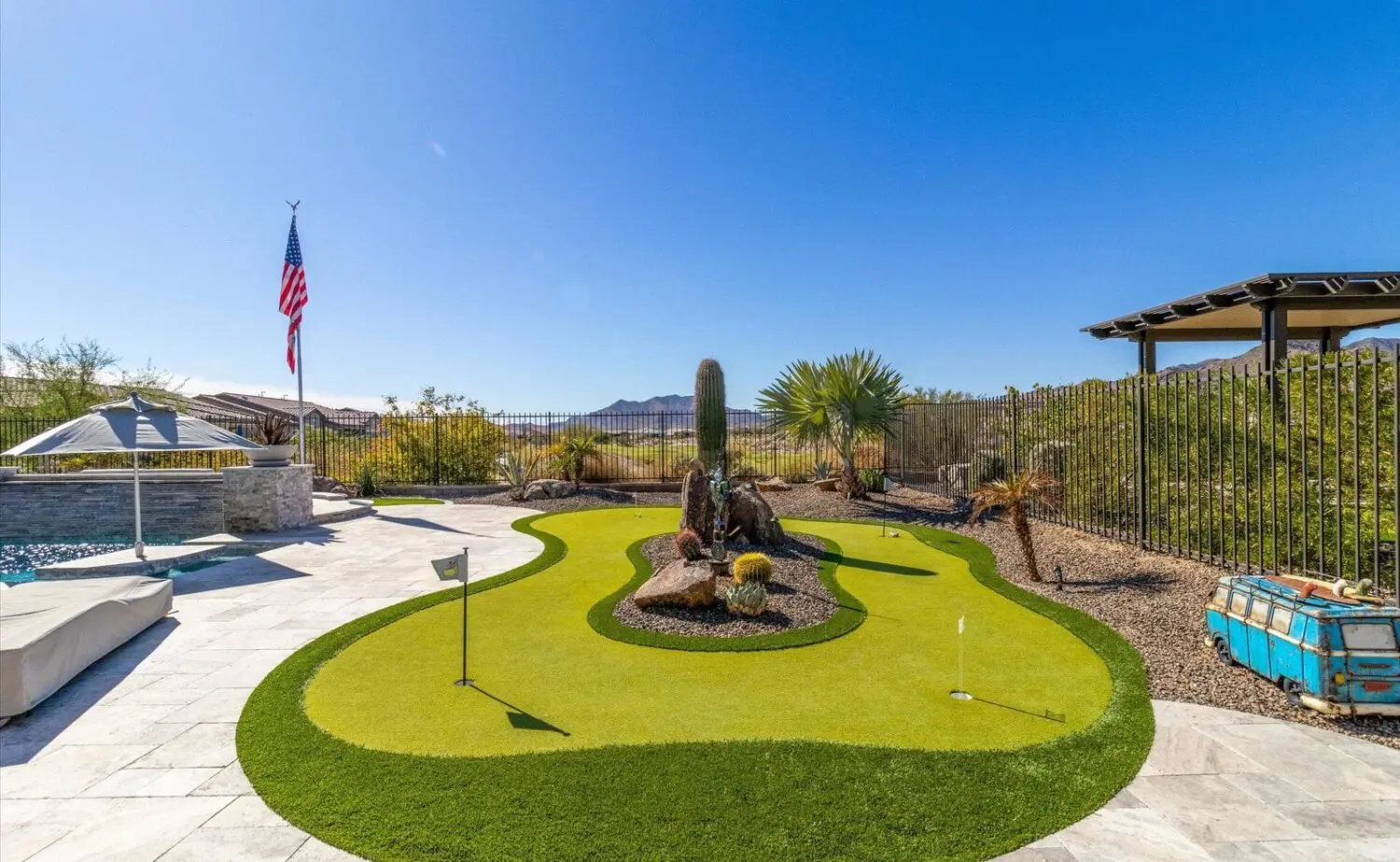 A backyard in Phoenix AZ features a small artificial putting green with turf installation, three golf holes, desert landscaping with a large cactus, a pool, an American flag, distant mountains, and a clear blue sky.