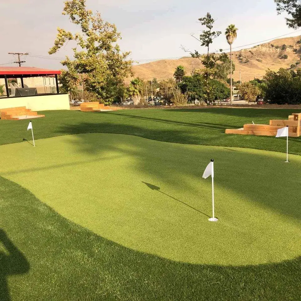 A small outdoor putting green with three white flags sits on premium artificial turf, surrounded by wooden benches, trees, and hills in the background under a partly cloudy sky—perfect for Paradise Valley AZ relaxation.