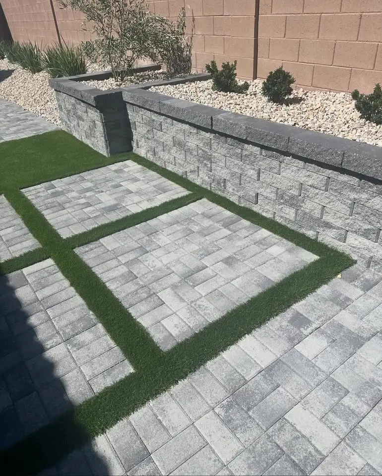 A backyard patio with gray stone pavers arranged in a grid pattern, separated by narrow strips of artificial grass, next to a low retaining wall and landscaped plants.