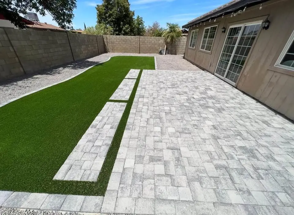 A backyard in Phoenix AZ with light gray stone pavers, a rectangular patch of artificial grass bordered by pavers, stone walls, and a beige house featuring large windows and glass doors.