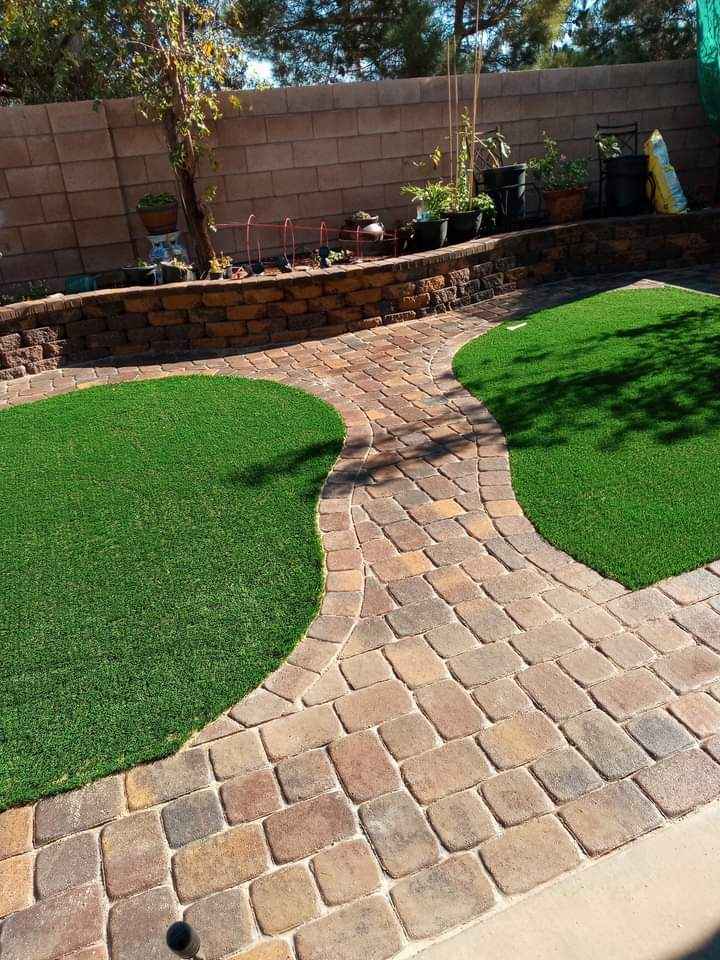 A backyard in Paradise Valley AZ with a curved stone walkway, bordered by artificial turf on both sides, brick retaining walls, and potted plants along a block wall under bright sunlight.